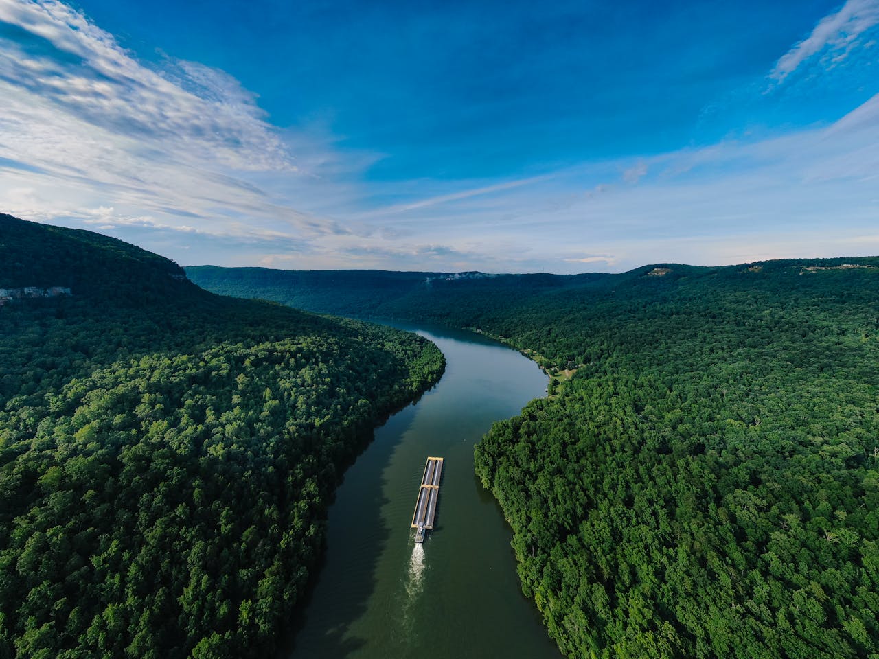 Aerial shot of a barge navigating a winding river through lush green forests under a clear blue sky.